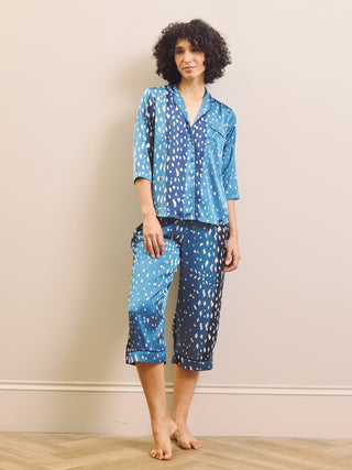 A woman modelling a two-piece capri pyjama set with a blue and white spotted pattern, consisting of a short-sleeve button-up shirt and matching cropped pants, standing against a beige wall.