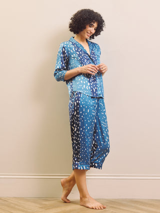 A woman modelling a matching two-piece pyjama set with a blue and white spotted pattern, standing barefoot against a plain beige wall.