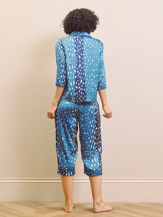 A woman viewed from behind wearing a matching two-piece capri pyjama set with a blue and white spotted pattern, standing barefoot on a light wood floor against a beige wall.