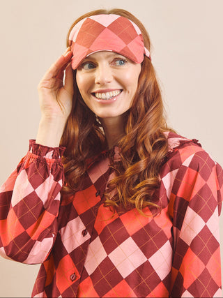 A smiling woman with long wavy red hair wears a matching argyle-patterned sleep mask and a matching long-sleeve top. The pattern features shades of red, pink, and orange on a light beige background, set against a solid light beige studio backdrop.