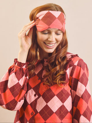 A smiling woman with long wavy red hair wears a matching argyle sleep mask and long-sleeve top in shades of red, pink, and burgundy against a plain beige background.