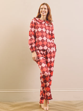 A woman modelling a matching two-piece pyjama set with a red, pink, and white harlequin diamond pattern, standing against a beige wall and light wood floor.