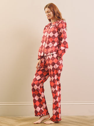 A woman modeling a two-piece pyjama set with a red, pink, and white harlequin diamond pattern, standing barefoot on a light wood floor against a beige wall.