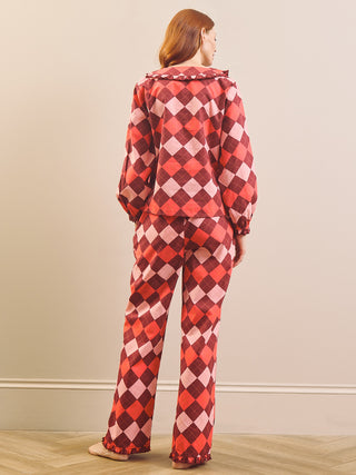 A woman stands with her back to the camera wearing a matching long-sleeve pyjama set with a red, pink, and burgundy harlequin diamond pattern, set against a neutral beige wall and light wood floor.