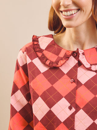 A close-up of a woman wearing a long-sleeve collared pyjama shirt with a pink, red, and orange argyle pattern and a ruffled Peter Pan collar.