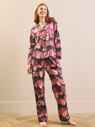 A model wearing a matching pyjama set with a pink and red fig pattern on a black background, set against a neutral beige wall and light wood floor.