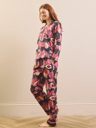 a model wearing a matching pyjama set with a pink and red fig pattern on a black background, set against a neutral beige wall and light wood floor.