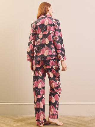 A woman stands with her back to the camera wearing a matching pyjama set with a pink and red fig pattern on a black background, set against a neutral beige wall and light wood floor.