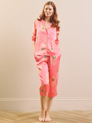Model wears satin capri pyjamas with a peacock feather print and a pink base against a beige background. 
