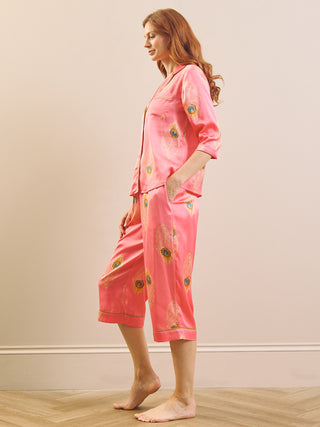 Model wears satin capri pyjamas with a peacock feather print and a pink base against a beige background. 