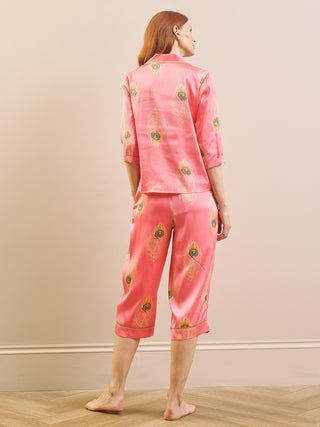 Model wears satin capri pyjamas with a peacock feather print and a pink base against a beige background. Back detail. 