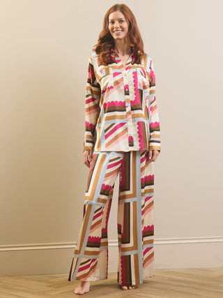 A woman modeling a matching two-piece pajama set with a retro geometric stripe pattern in pink, brown, beige, and light blue on a cream background, standing against a beige wall.