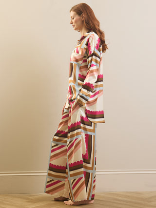 A woman stands barefoot in profile wearing a long-sleeve, wide-leg patterned robe or pajama set with geometric stripes in pink, brown, beige, and light blue on an off-white base, against a plain beige wall and light wood floor.