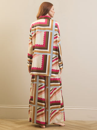 A woman stands with her back to the camera wearing a matching long-sleeve top and wide-leg skirt set in a cream base with geometric block patterns in pink, brown, beige, and light blue.