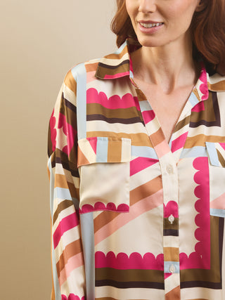 A close-up of a woman wearing a long-sleeve button-up pyjama shirt with a retro striped pattern in pink, brown, beige, and light blue. The shirt features scalloped edges along the pocket flaps and collar.