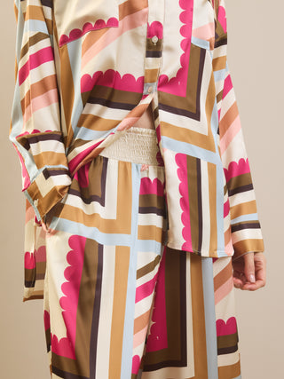 A close-up of a person wearing a matching long-sleeve button-front pyjama top and skirt set with a bold geometric pattern in beige, brown, pink, light blue, and white.