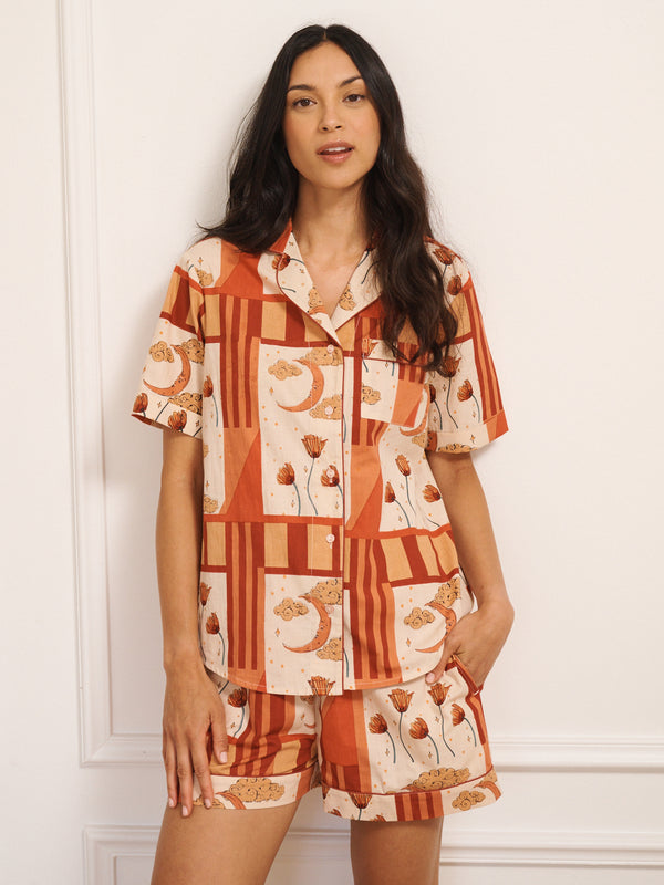 Women’s cotton shortie pyjamas in a natural patchwork sky and flower print, featuring a short-sleeve button-through shirt and matching shorts, worn by model standing against a white panelled wall.
