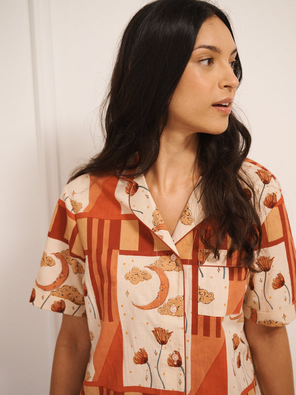 Model wears Women’s cotton shortie pyjamas in a natural patchwork sky and flower print, featuring a short-sleeve button-through shirt and matching shorts, worn by model standing against a white panelled wall.