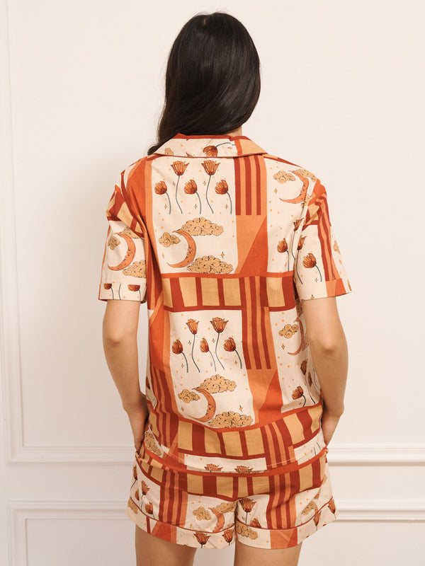 Model wears Women’s cotton shortie pyjamas in a natural patchwork sky and flower print, featuring a short-sleeve button-through shirt and matching shorts, worn by model standing against a white panelled wall. Back view. 