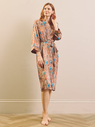 A woman stands barefoot in a room with a beige wall and light wood floor, wearing a knee-length feather patterned dressing gown robe with three-quarter sleeves and a self-tie belt.