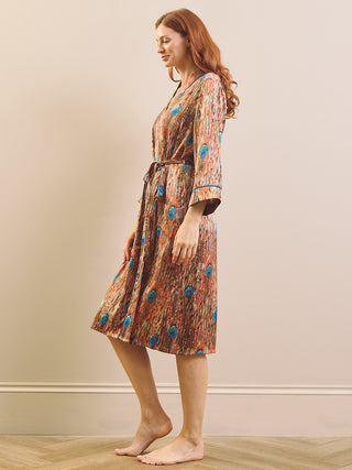 A woman with red hair stands barefoot in profile wearing a knee-length patterned dressing gown robe with three-quarter sleeves and a tie belt. The robe has an abstract multicolor print with brown, orange, and blue tones.