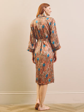 A woman stands barefoot indoors wearing a knee-length patterned dressing gown robe with a tie waist and three-quarter sleeves, shown from the back against a plain beige wall.