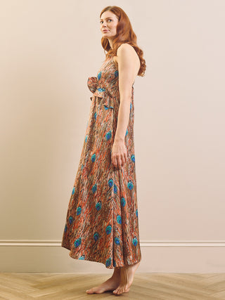 A woman stands barefoot in profile wearing a sleeveless maxi nightdress with a brown abstract pattern and scattered blue and turquoise circular motifs, set against a plain beige wall and light wood floor.