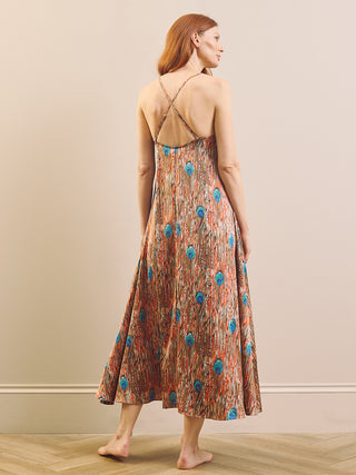 A woman stands barefoot indoors wearing a sleeveless maxi nightdress shown from the back. The dress has thin crisscross straps at the open back and an allover abstract print in rust orange, beige, and bright blue.