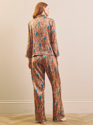 A woman stands with her back to the camera wearing a matching two-piece pyjama set with a brown and orange abstract feather pattern and blue circular accents, set against a neutral beige wall and light wood floor.