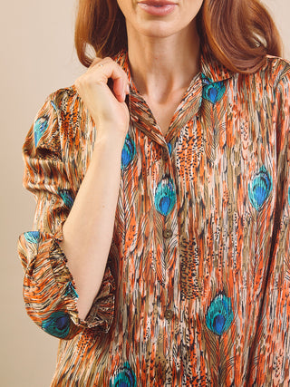 A close-up of a woman wearing a long-sleeve button-up pyjama shirt with an orange, brown, and black feather print and bright blue eye spots, against a plain beige background.