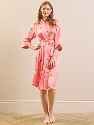 A woman modelling a pink satin dressing gown robe with gold and green floral patterns, standing barefoot against a plain beige wall and light wood floor.