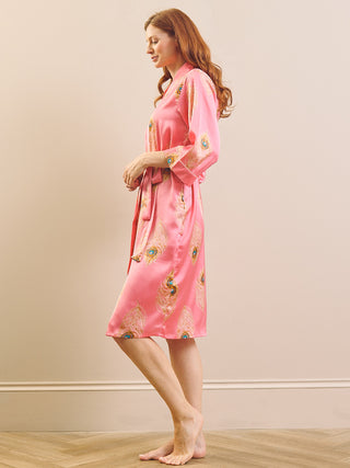 A woman wearing a pink satin robe with peacock feather patterns, standing barefoot in a studio setting with a beige wall and light wood floor.