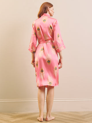 A woman stands barefoot indoors wearing a pink satin-like robe with gold-toned peacock feather motifs, shown from the back against a plain beige wall and light wood floor.