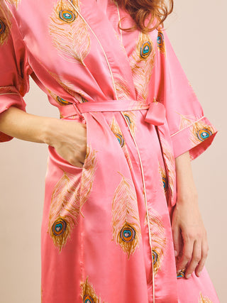 A close-up of a woman wearing a pink satin robe with a peacock feather print and a matching tie belt, shown against a plain beige background.