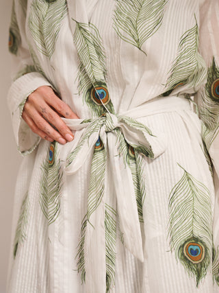 Women’s white cotton seersucker mini robe with green peacock feather print and tie waist, worn by model standing barefoot against a white panelled wall. Tie detail. 