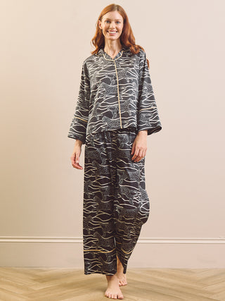 A woman stands barefoot in a studio setting wearing a matching two-piece pyjama set with a black base and an abstract wavy line pattern in white and gold including a Cheetah. The set includes a long-sleeve button-front top and matching loose pants.
