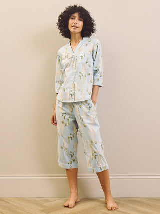 Woman wearing a light blue floral wisteria capri pyjama set against a beige wall.