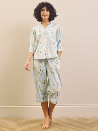 Woman wearing a light blue floral wisteria capri pyjama set against a beige wall.