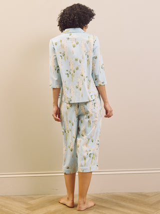 Woman wearing a light blue floral wisteria capri pyjama set against a beige wall.