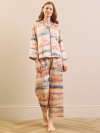 A woman stands barefoot in a bedroom setting wearing a two-piece pyjama set with a watercolour-style landscape print in soft pink, blue, and beige tones with dark piping.