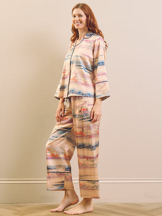 A woman modeling a two-piece pyjama set with a colorful abstract landscape print, featuring a long-sleeve button-up top and matching wide-leg pants, set against a neutral beige wall.