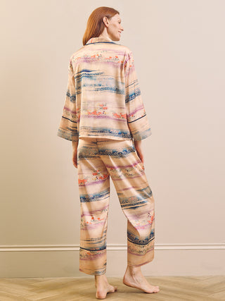A woman stands barefoot in profile wearing a matching two-piece pyjama set with a soft abstract landscape print in beige, pink, blue, and brown tones, photographed against a neutral beige wall and light wood floor.