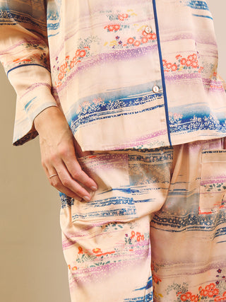 A close-up of a person wearing a matching pyjama set with a soft pastel abstract floral print. The pajamas feature a light beige base with blue, pink, and orange watercolor-style stripes and floral motifs, accented by dark blue piping.