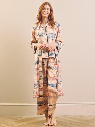 A woman stands barefoot in a bedroom setting wearing a matching two-piece pyjama set consisting of a long-sleeve button-up shirt and a long dressing gown robe, both in a soft beige base fabric with a multicolour landscape print of blue, pink, and orange.