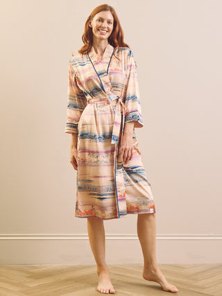 A woman stands barefoot in a studio wearing a long wrap dressing gown robe with a multicolour abstract pattern on a light beige base. The robe has blue, pink, orange, and purple horizontal stripe-like bands.
