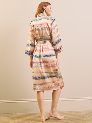 A woman stands barefoot indoors wearing a knee-length satin dressing gown robe with a tie waist and three-quarter sleeves. The robe has a beige base with horizontal multicolor stripes in blue, pink, and orange, shown from a rear three-quarter view against a plain beige wall and light wood floor.
