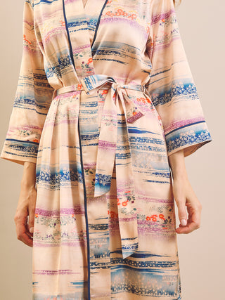 A close-up of a person wearing a long dressing gown robe with a tie waist and wide sleeves. The robe has a soft beige base with horizontal multicolor patterns in blue, pink, purple, and orange, plus dark blue piping along the edges.