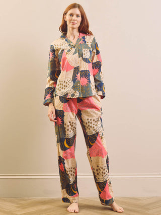 A woman stands barefoot in a room wearing a matching two-piece pyjama set with a colourful abstract patchwork print. The set includes a long-sleeve button-front top and matching pants.