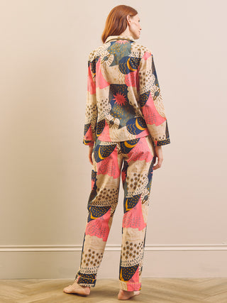 A woman stands barefoot in a room wearing a matching long-sleeve pyjama set with a colourful abstract patchwork print, photographed from the back.