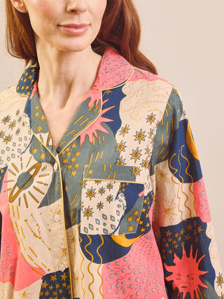 A close-up of a woman wearing a long-sleeve button-up pyjama shirt with a colourful patchwork print featuring planets, stars, suns, and abstract shapes in pink, navy, beige, and mustard yellow.